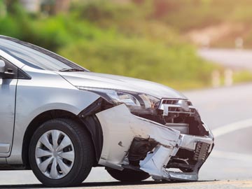 Ein silbernes Auto mit beschädigtem vorderen Stoßfänger ist am Straßenrand geparkt, vor einem unscharfen grünen Hintergrund.